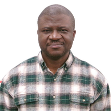 Black man in front of white background, wearing plaid shirt